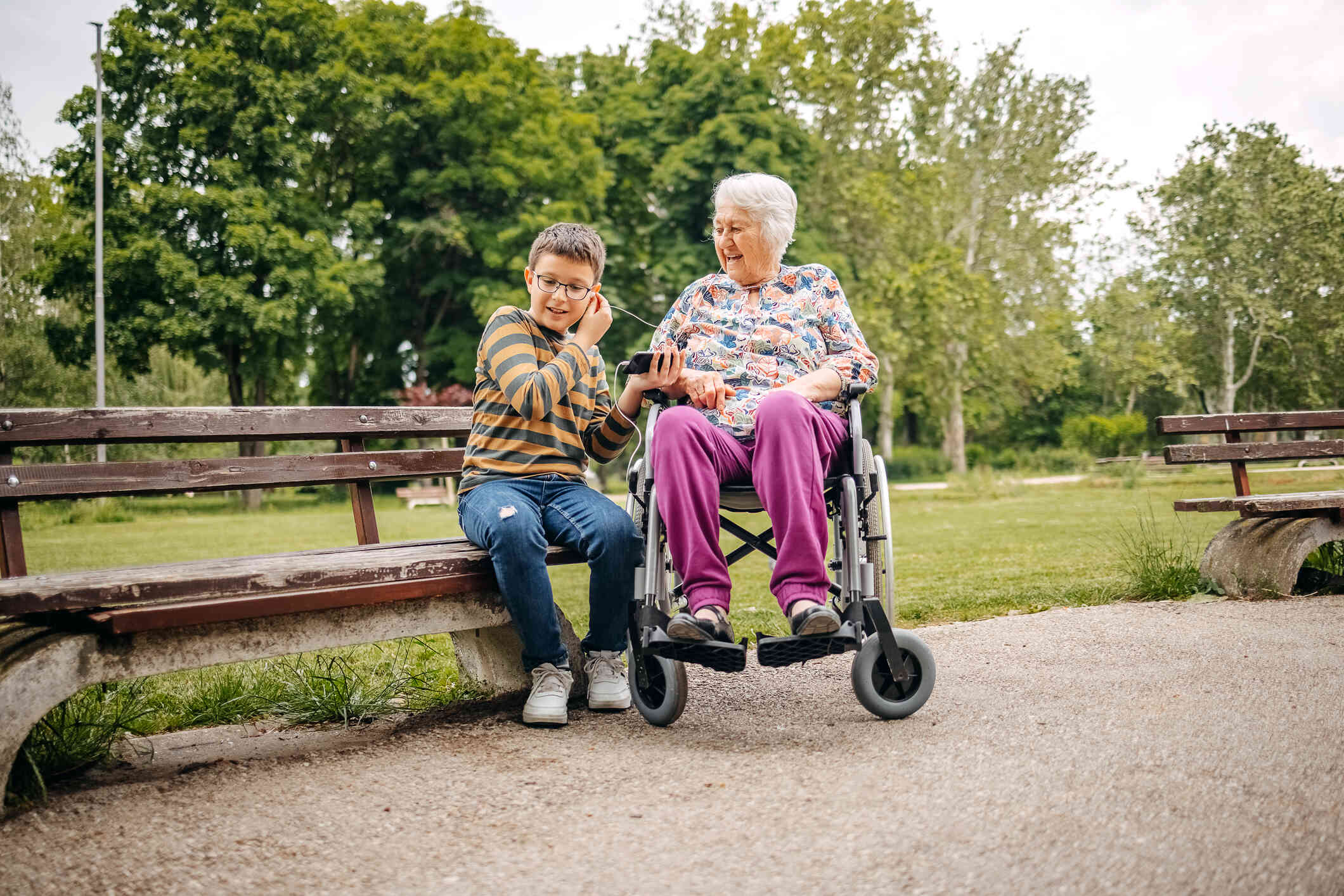 An elderly woman in a wheelchiar sits outside next to a park bench with her grandson sitting next to her at they share headphones and listen to music together.